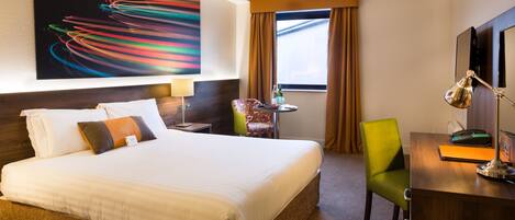 A hotel room with a large bed, a desk with a lamp, a chair, a window with curtains, and a colorful abstract painting on the wall.