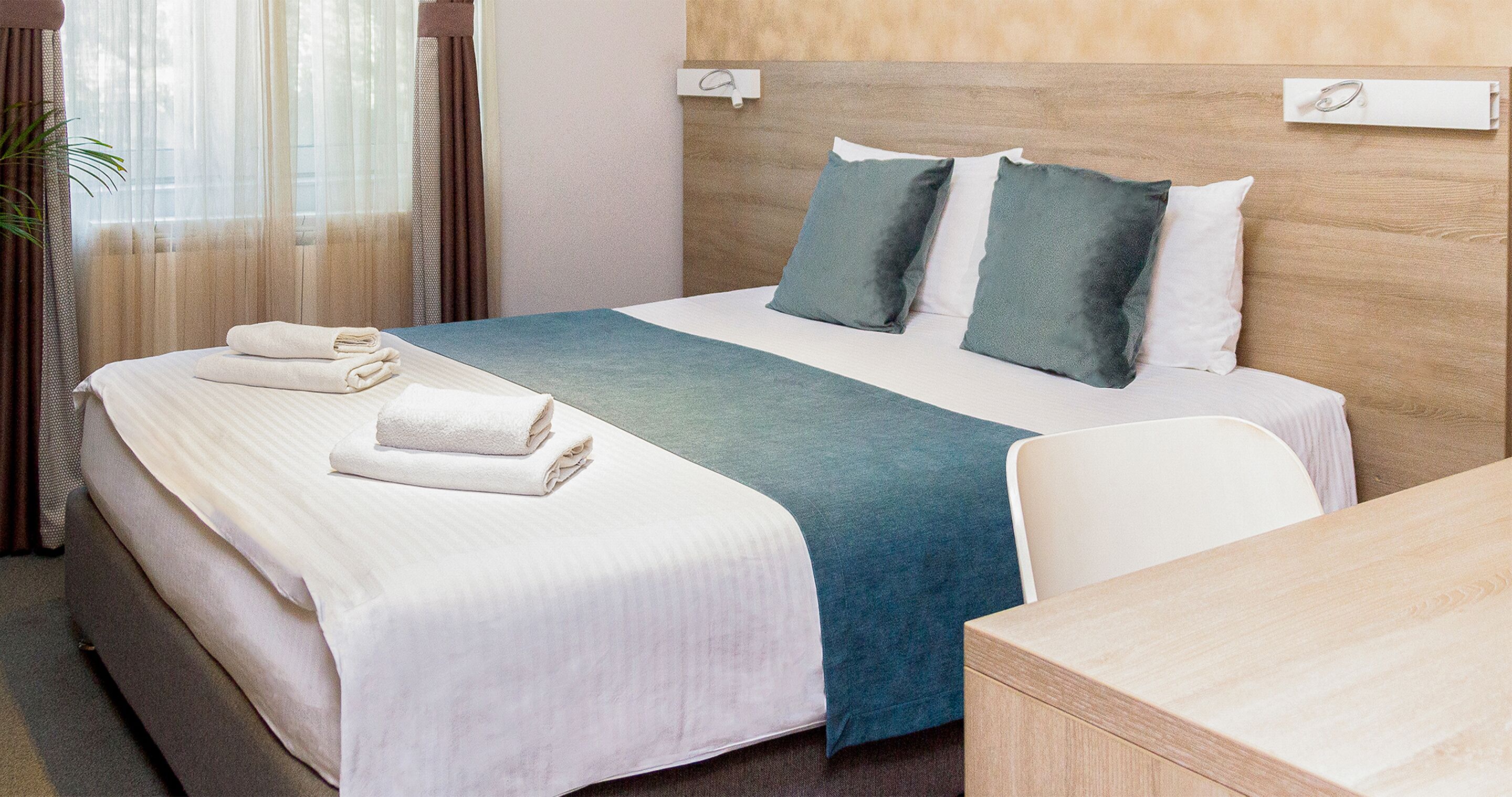 A neatly made bed with white linens and a teal blanket, a wooden headboard, and a small wooden desk.