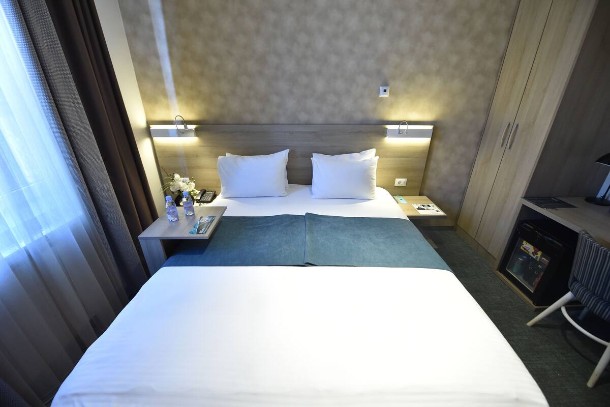 A hotel room with a bed, two bedside tables, a nightstand, a lamp, a chair, and a small refrigerator.