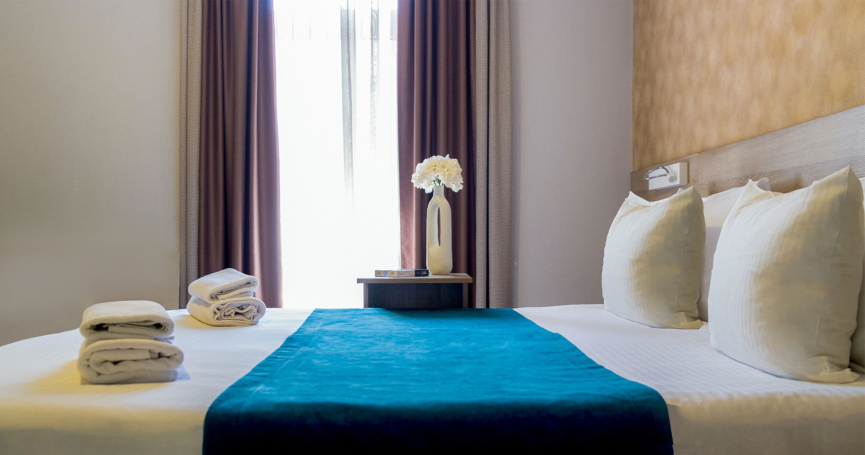 Una cama bien hecha con una colcha azul, almohadas blancas y un jarrón con flores.