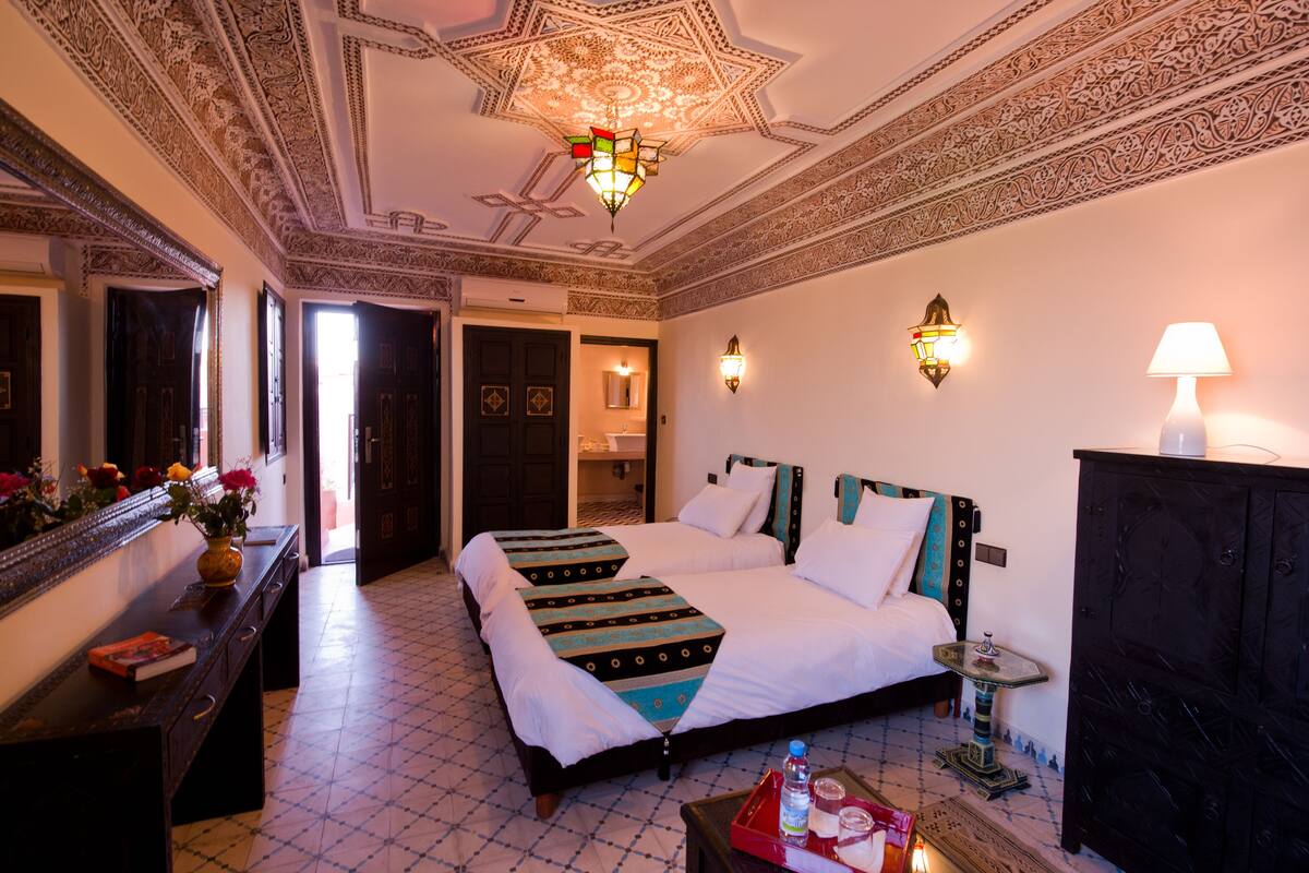 A hotel room with two beds, a dresser, a mirror, a chandelier, and decorative wall sconces.