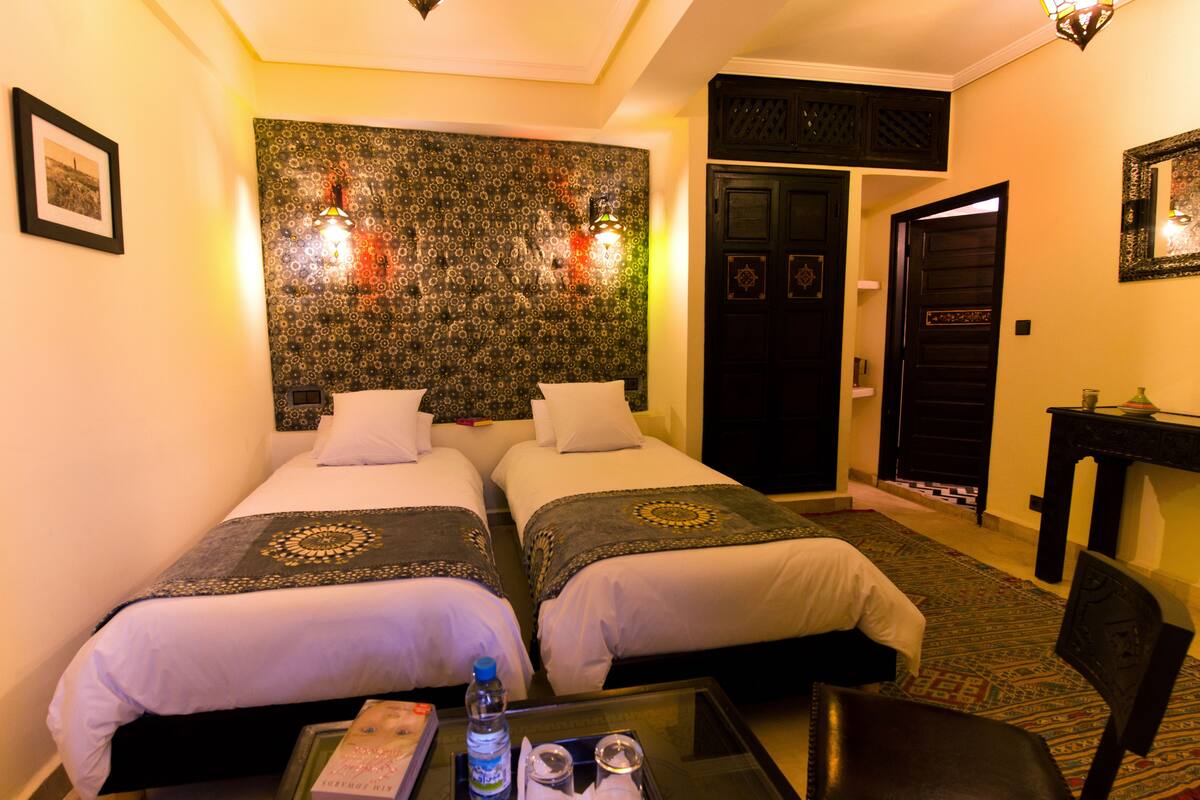 A hotel room with two beds, a small table with a bottle and glasses, a chair, and a wall tapestry.