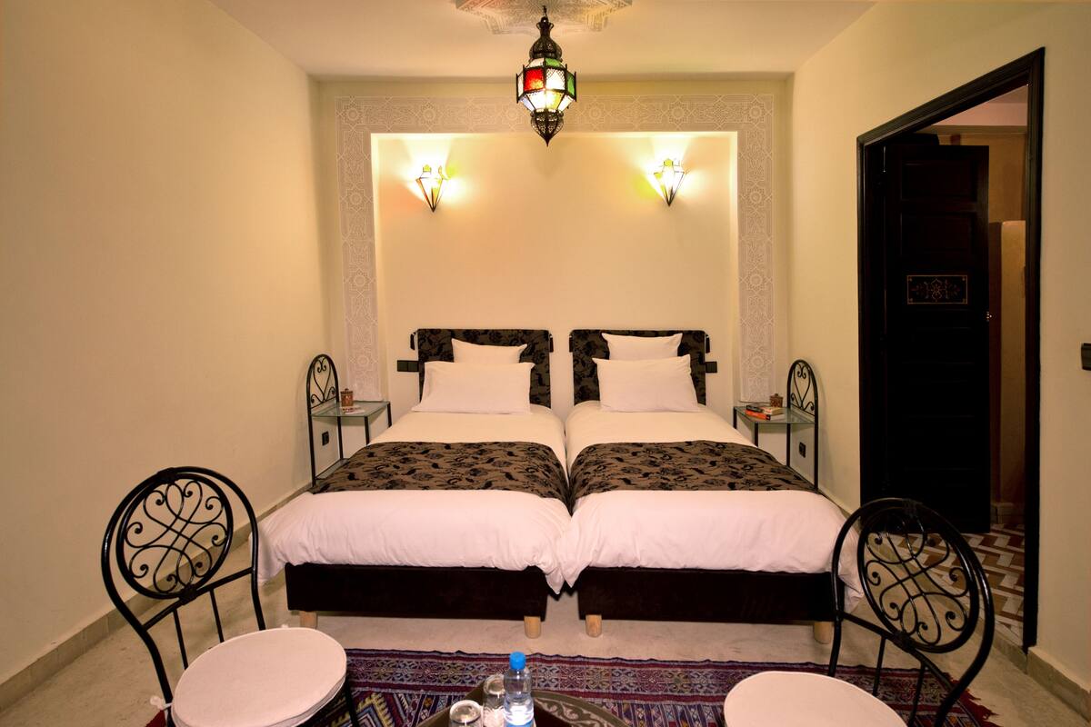 A double bed with a patterned headboard, two bedside tables, two chairs, a patterned rug, and a decorative ceiling light.