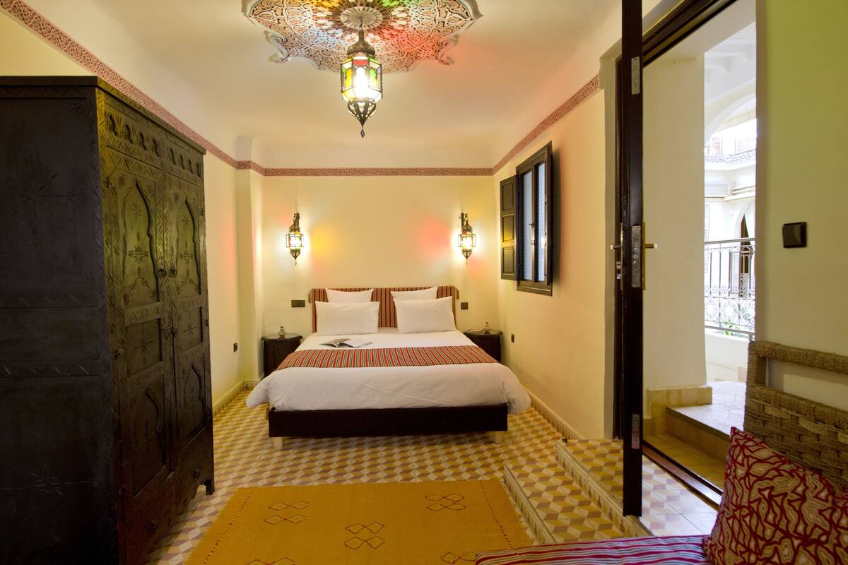 A hotel room with a large bed, two wall-mounted lamps, a decorative ceiling light, a checkered floor, and a view of the outside balcony.