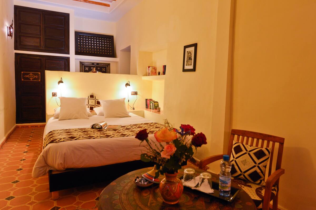 A hotel room with a bed, bedside table, and a chair. There is a vase with flowers on the table.