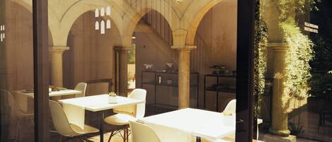 A glass-enclosed dining area with white tables and chairs, stone arches, and a view of the outdoor garden.