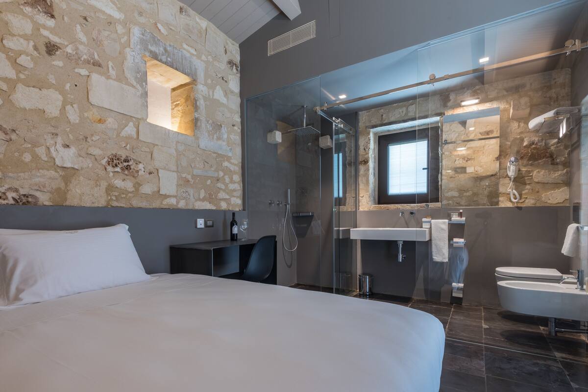 Une chambre d’hôtel moderne avec un mur en pierre, un grand lit, une douche en verre et une salle de bain équipée d’un lavabo et de toilettes.
