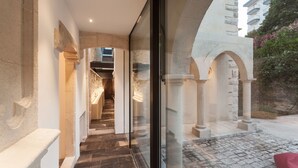 Un couloir bordé de colonnes en pierre et doté d’une porte en verre qui donne sur une cour dallée, où se trouve un coussin rouge.