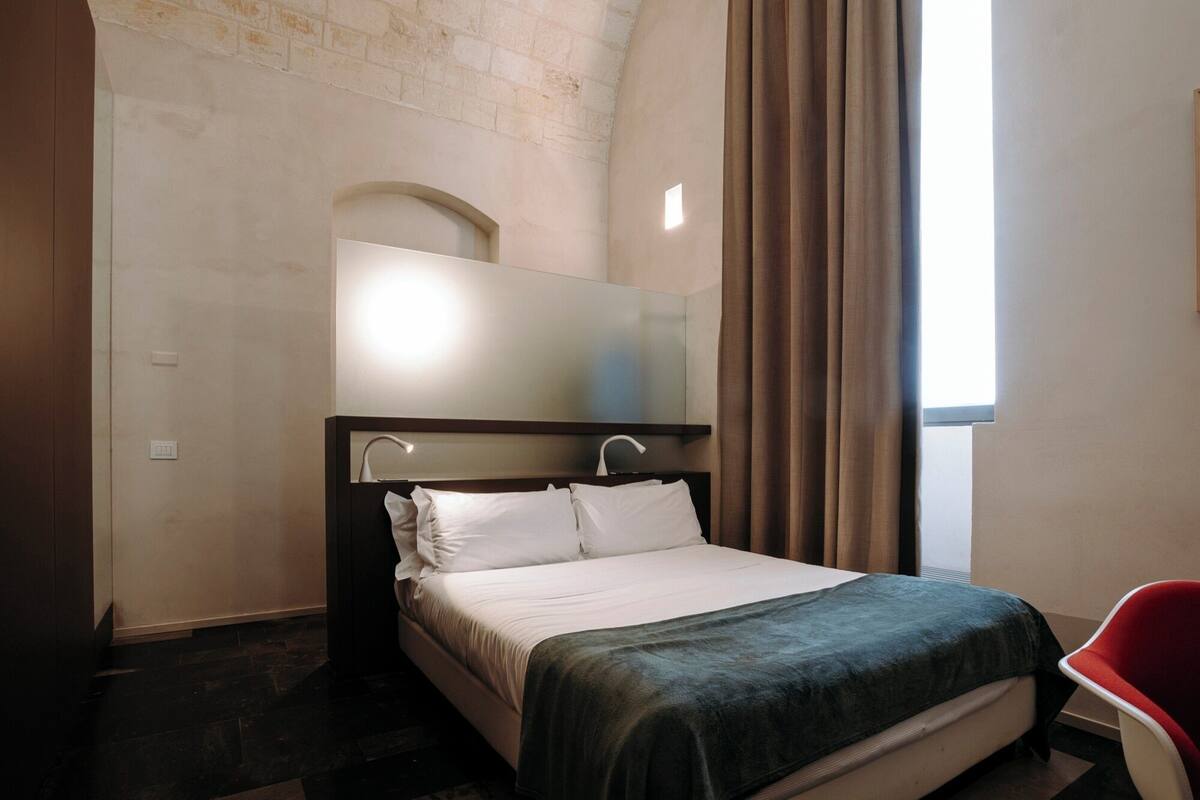 Une chambre d’hôtel avec un lit, un miroir, une fenêtre avec des rideaux et une chaise rouge.