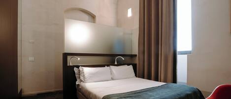 Une chambre d’hôtel avec un lit, un miroir, une fenêtre avec des rideaux et une chaise rouge.