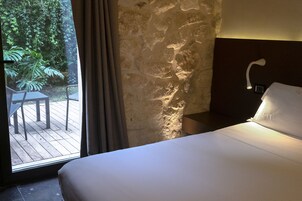 Habitación de hotel con cama, mesita de noche, lámpara de pared y vistas a una terraza de madera y follaje exterior.