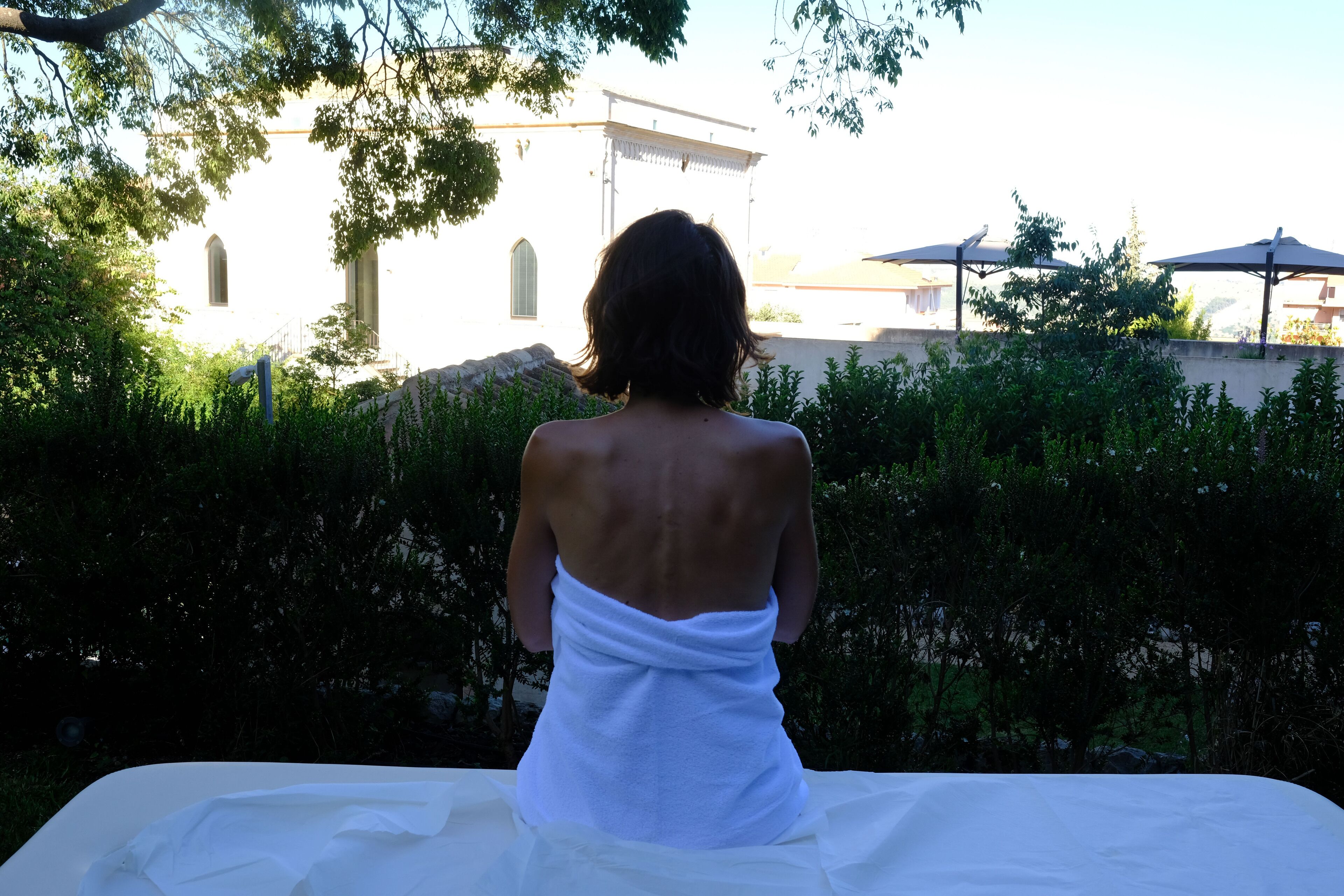 A person sitting on a bed with a white sheet, facing a building with arched windows and a white facade, surrounded by greenery.