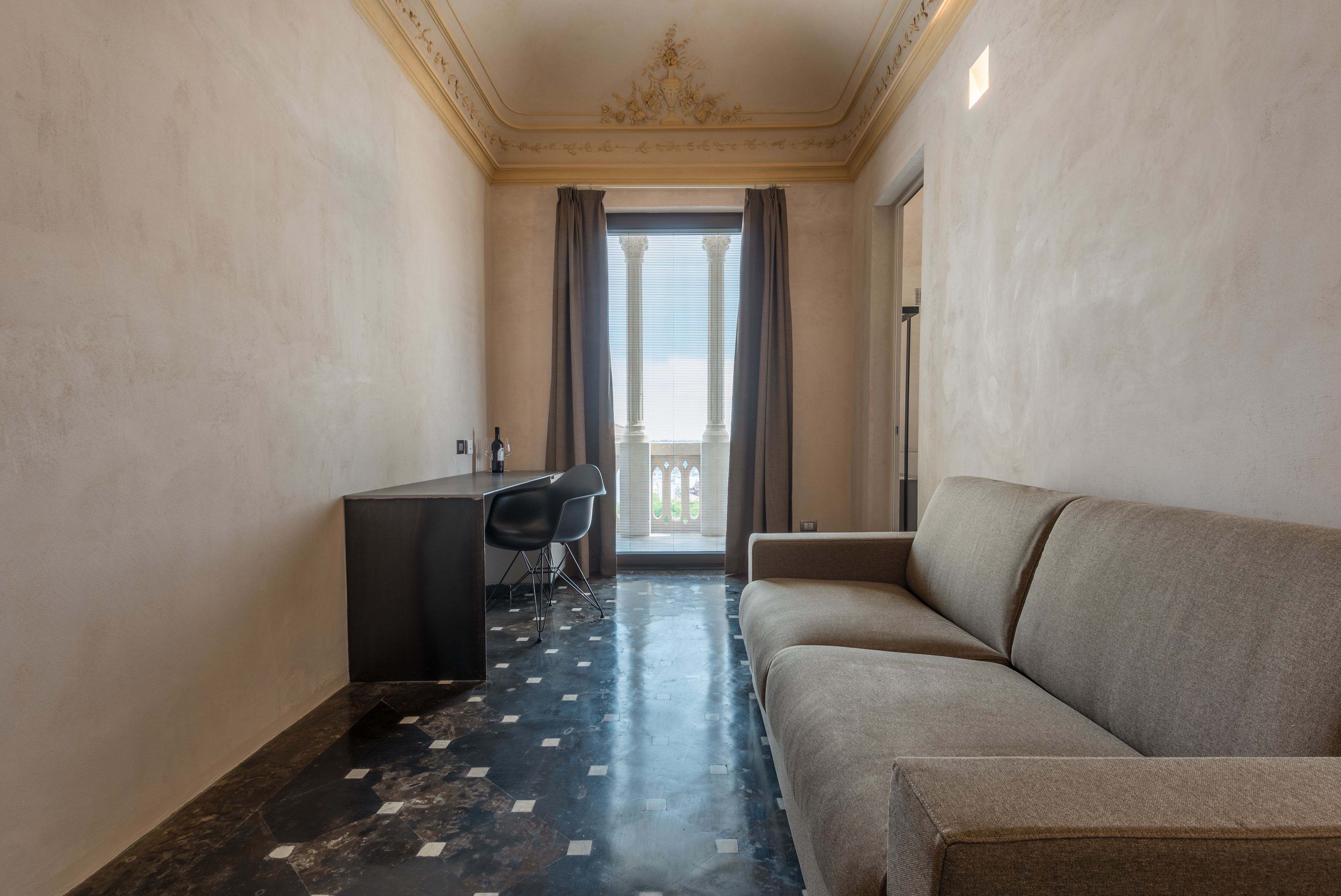 A hotel room with a sofa, a desk, and a chair. There is a view of a balcony with curtains.
