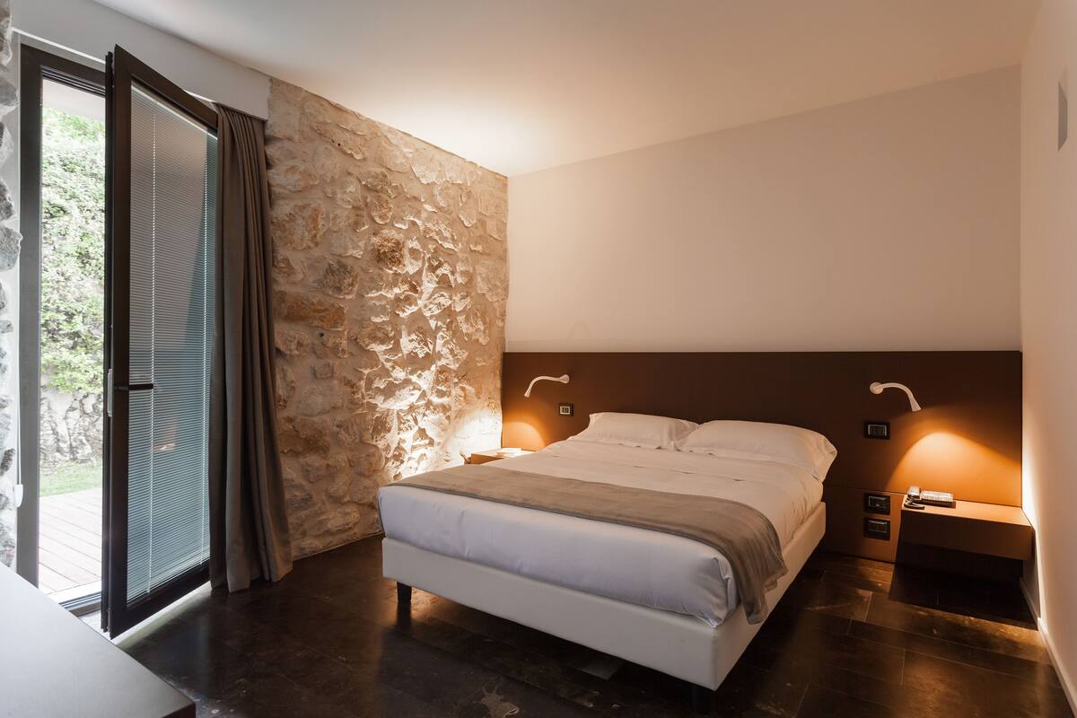 Une chambre avec un mur en pierre, un lit recouvert de draps blancs, une table de chevet avec une lampe et une porte-fenêtre coulissante donnant sur un balcon.