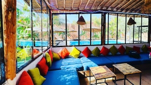 Una habitación con un sofá azul largo, cojines coloridos, una mesita de madera y grandes ventanales con vistas a una piscina y vegetación.