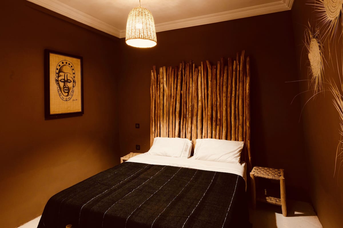 A bedroom with a bed, a hanging light fixture, a wooden headboard, a framed artwork, and a small stool.
