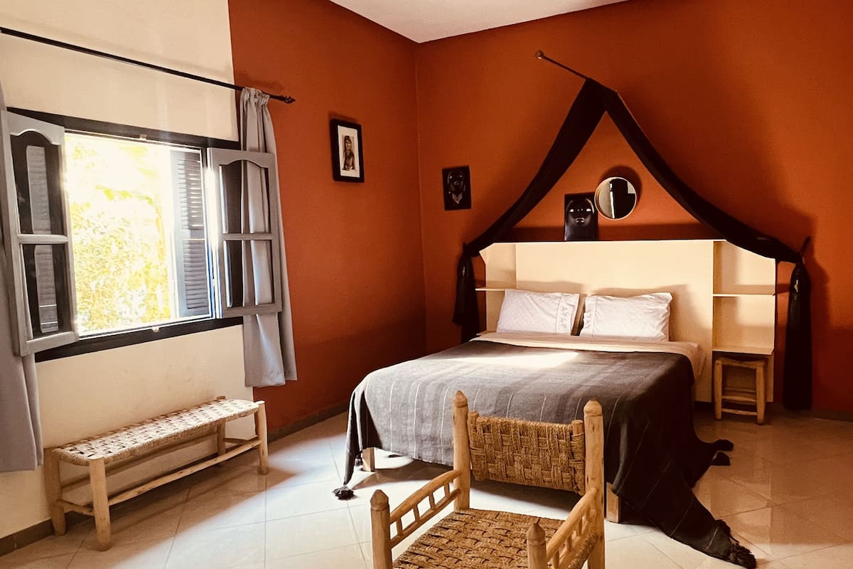 A bedroom with a bed, a bench, a chair, a window with curtains, and a wall with framed pictures.