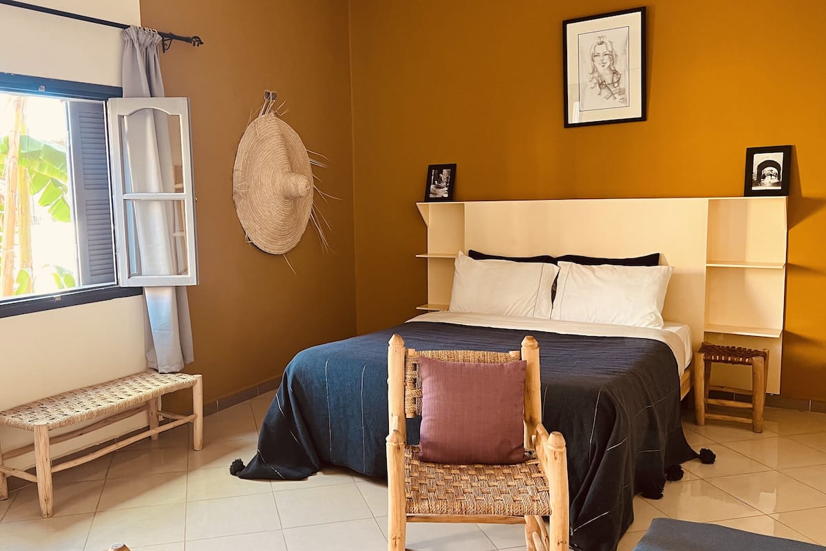 A bedroom with a bed, a chair, a bedside table, a window with curtains, and wall art.