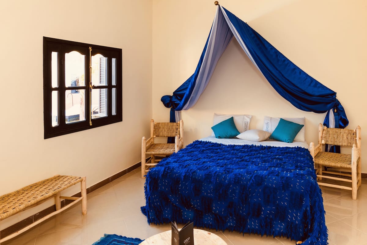 A bedroom with a bed, two chairs, a bench, a window, and a curtain.