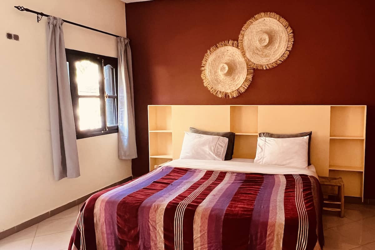 A bedroom with a bed, a window with curtains, a headboard with shelves, and two wall-mounted decorative items.
