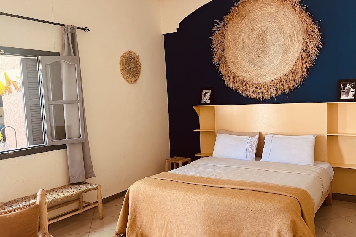 A bedroom with a bed, bedside tables, a window with shutters, a woven wall hanging, and a small stool.