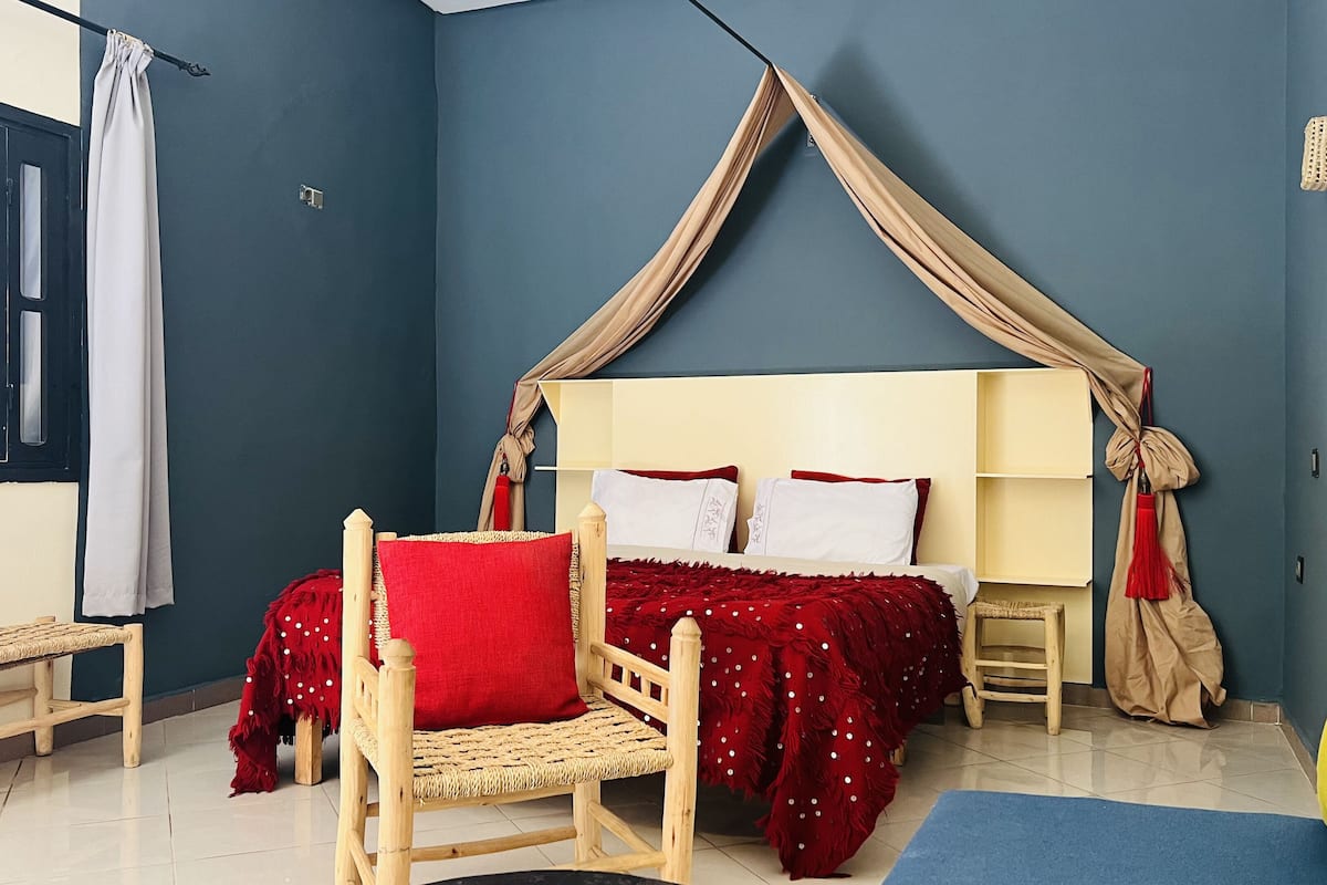 A bedroom with a canopy bed, a wooden chair, a small bedside table, and a window with blinds.