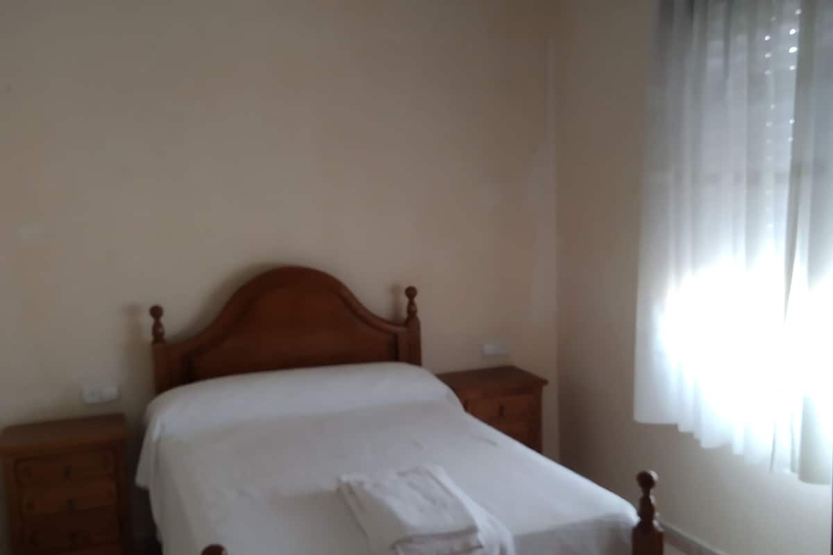 A bedroom with a wooden bed, white linens, a nightstand, and a window with curtains.