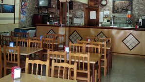 A restaurant with wooden tables and chairs, a counter with a cash register, and a stone wall with a clock and artwork. - Hostal El Viejo Galeón (Baiona)
