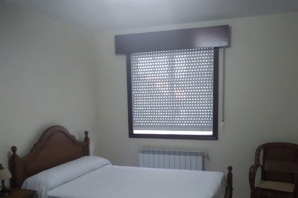 A room with two single beds, a wooden chair, a radiator, and a window with a metal screen.