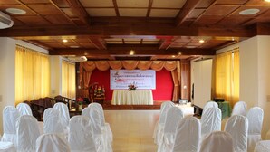 A conference room with rows of chairs covered in white cloth, a stage with a banner, and wooden ceiling beams.