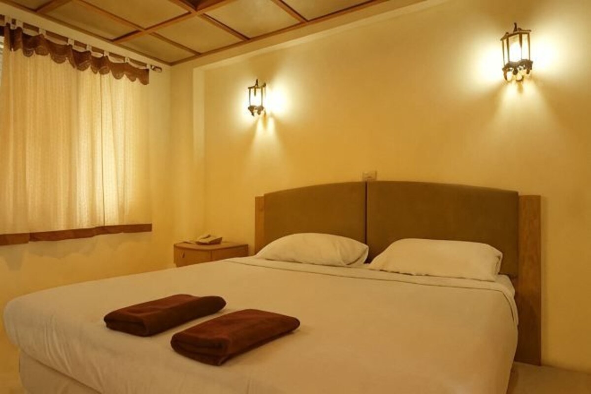 A hotel room with a large bed, two pillows, two towels, a nightstand, and two wall-mounted lights.