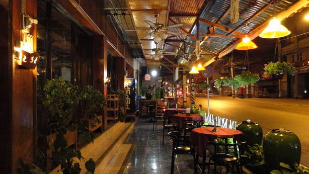 An outdoor cafe with tables and chairs, potted plants, and hanging lights.