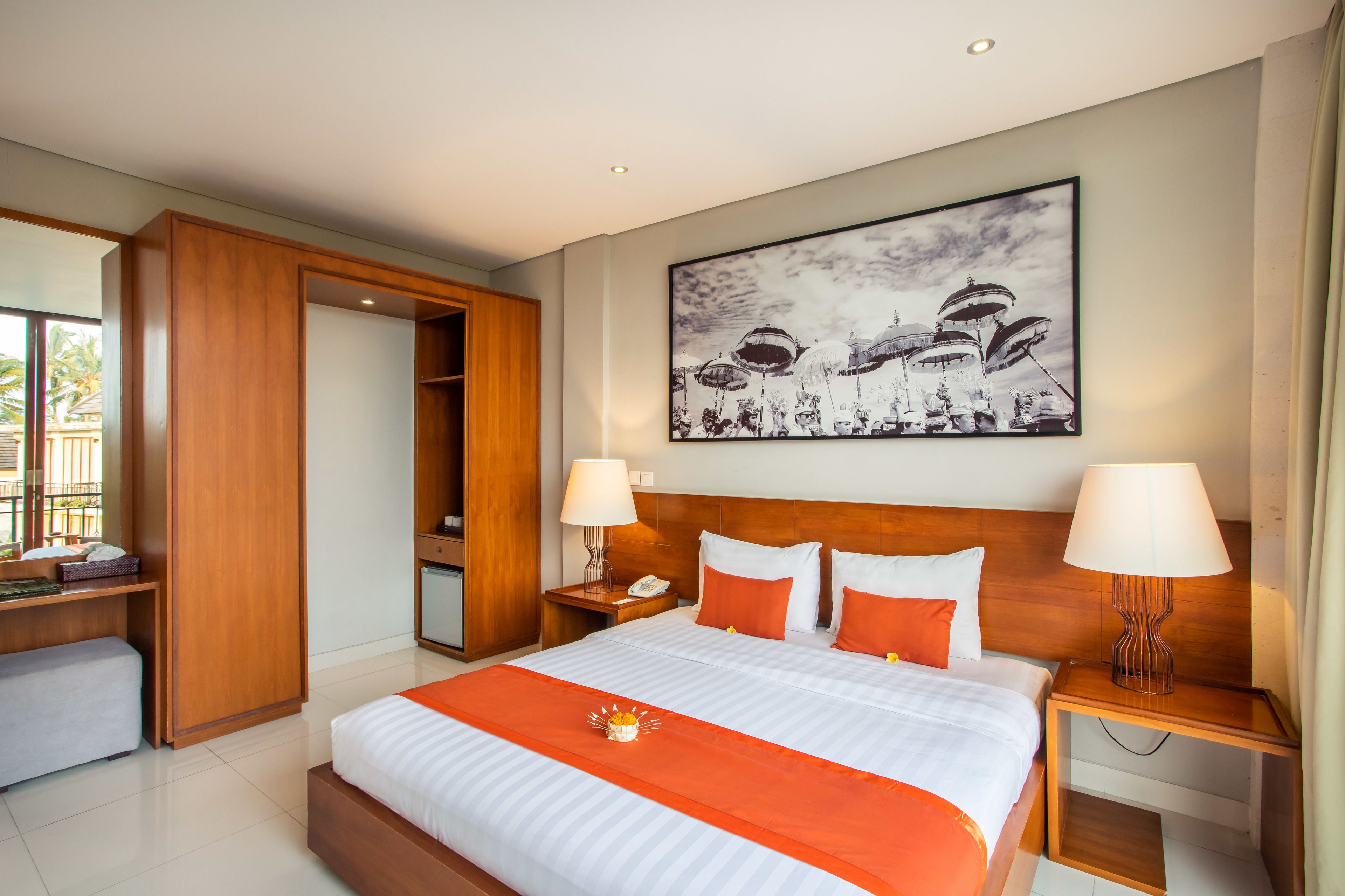 A hotel room with a large bed, two bedside tables with lamps, a wooden wardrobe, a desk with a chair, and a large framed artwork on the wall.