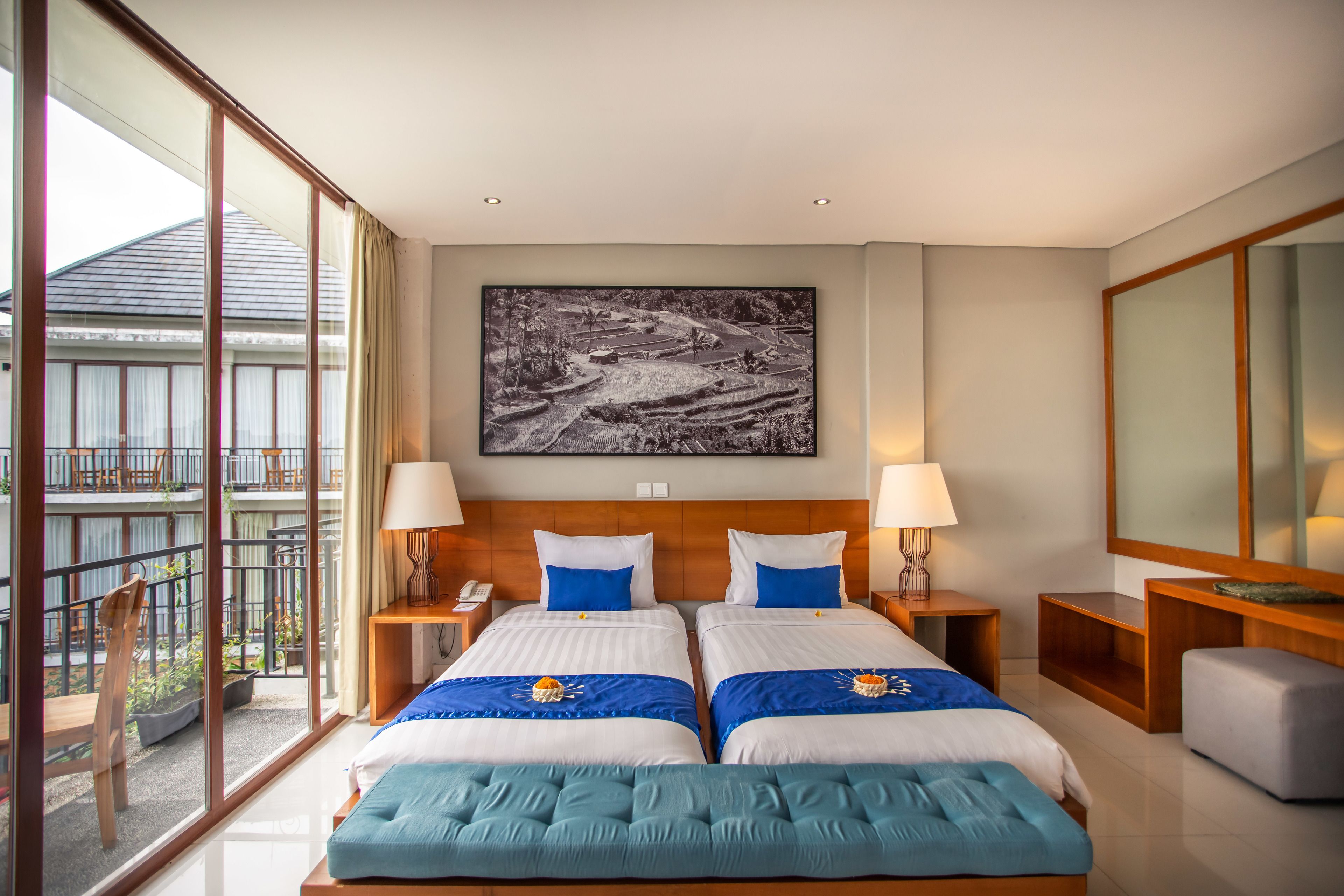 A hotel room with two beds, a large window, a balcony, and a painting on the wall.