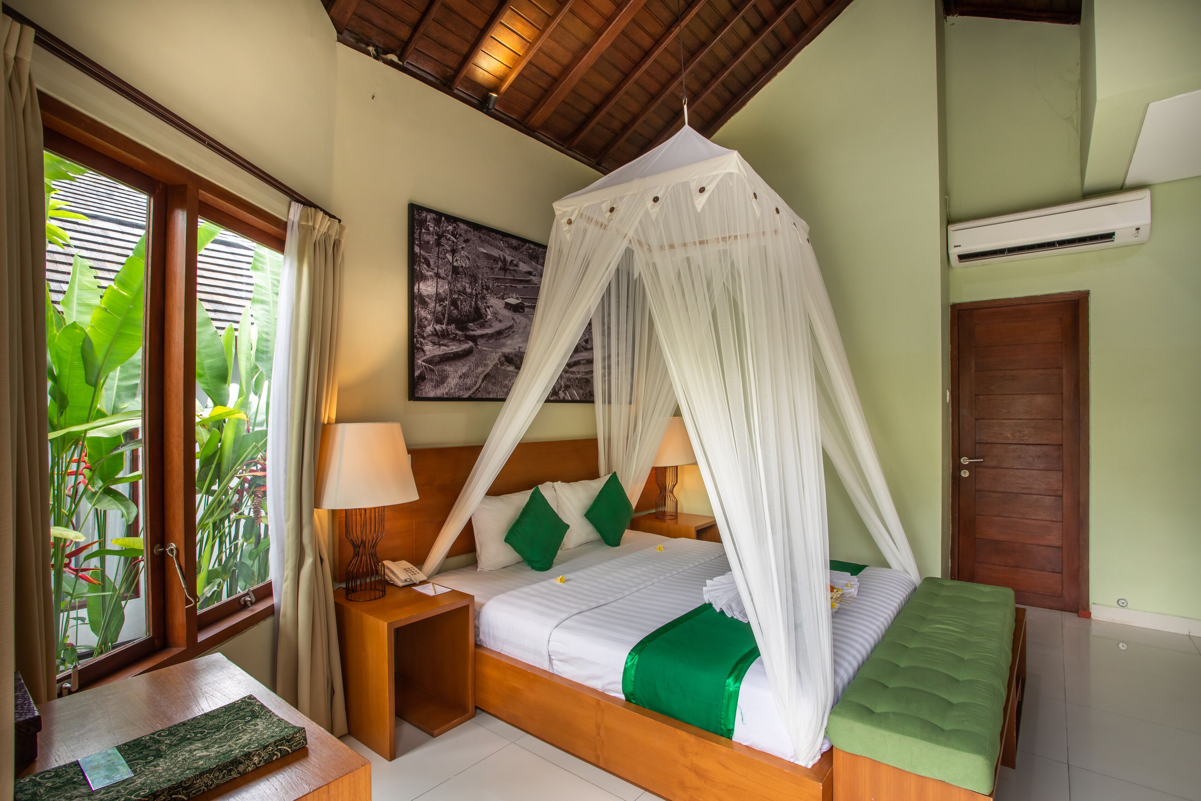 A bedroom with a canopy bed, a bedside table with a lamp, a green throw blanket, and a view of greenery through the window.