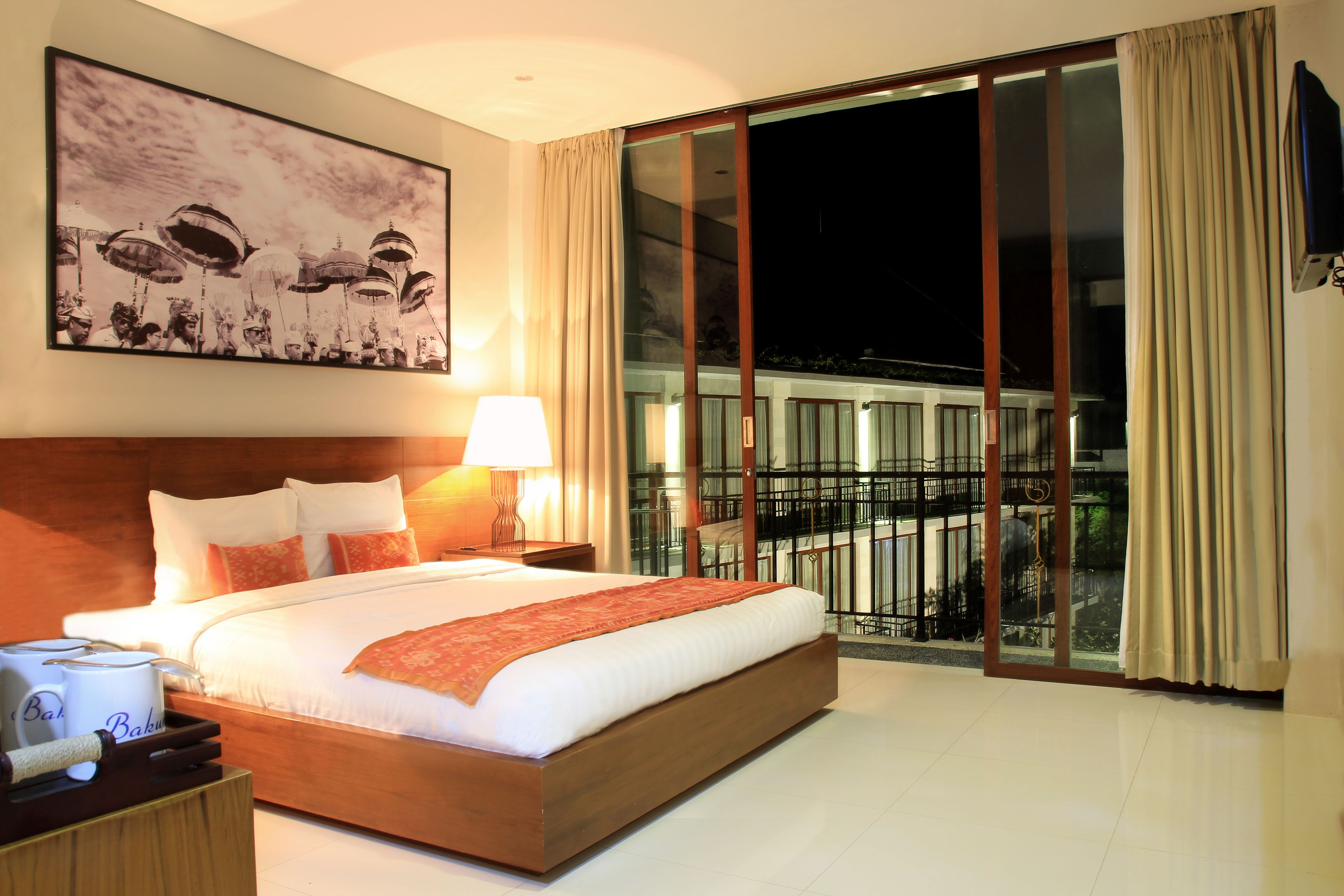 A hotel room with a large bed, a nightstand with a lamp, a painting on the wall, and a balcony with a railing.
