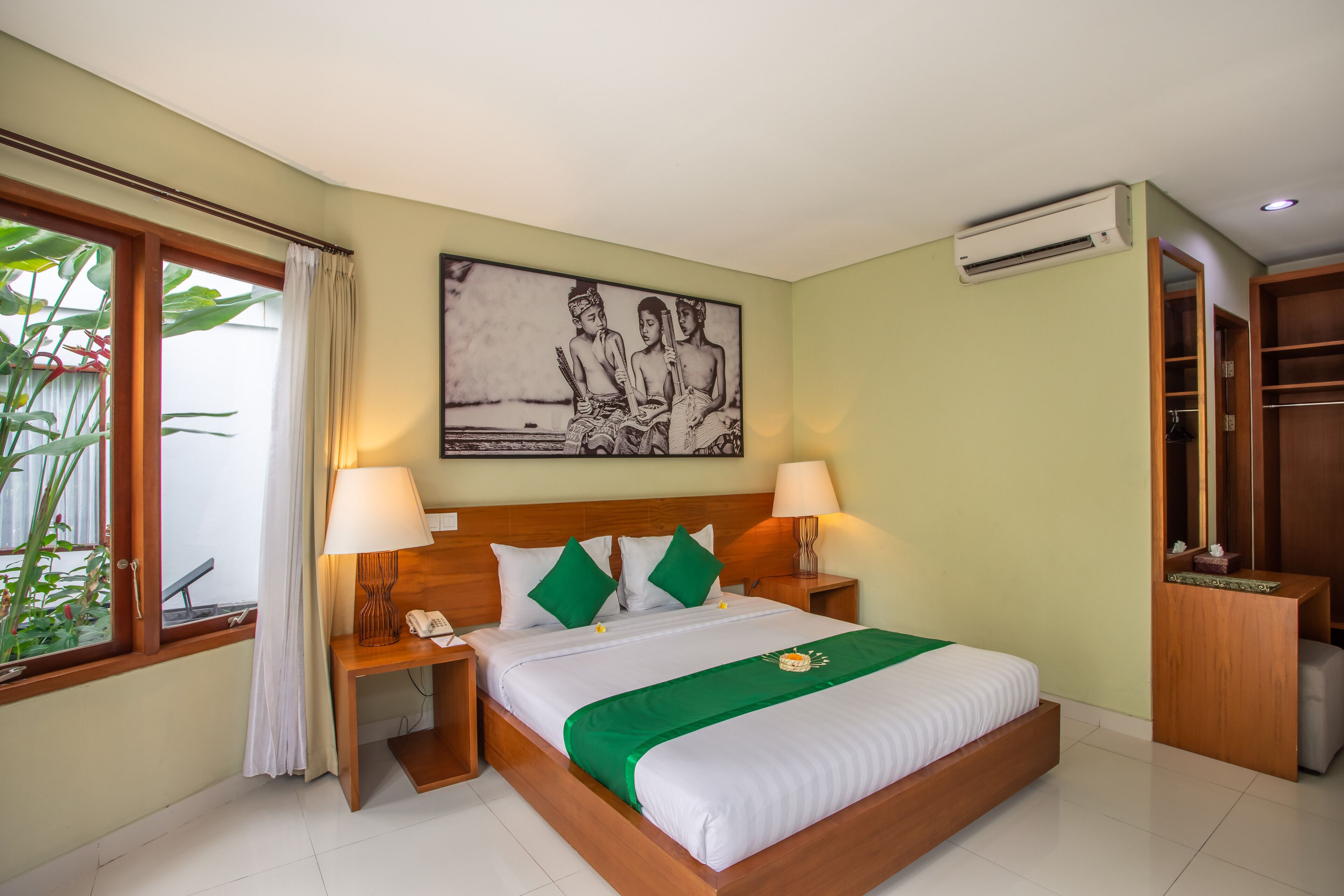 A hotel room with a bed, two bedside tables, a nightstand, a window with curtains, an air conditioning unit, and a wooden wardrobe.