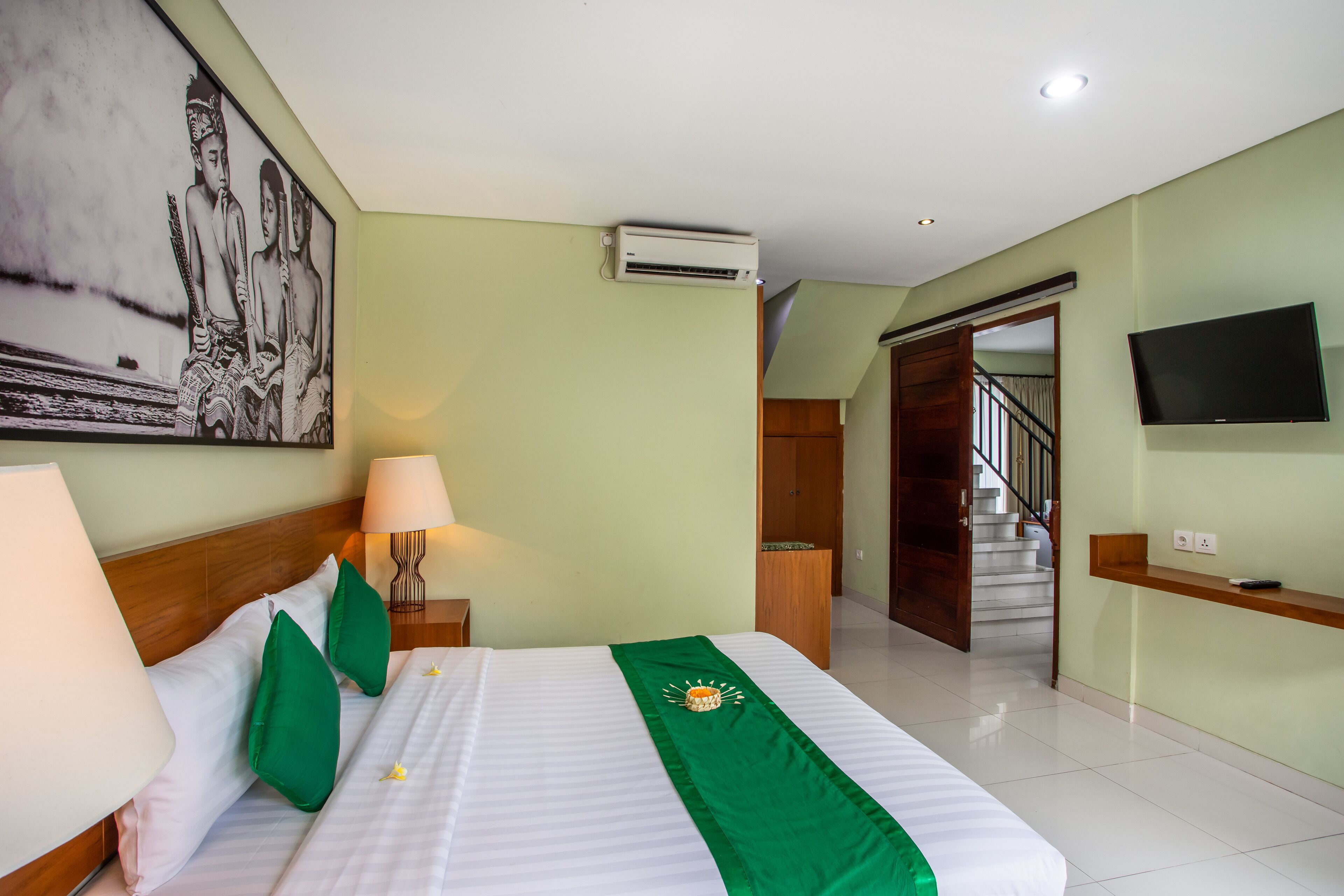 Habitación de hotel con cama, almohadas verdes, mesita de noche con lámpara, televisor montado en la pared y una escalera que sube a otro piso.