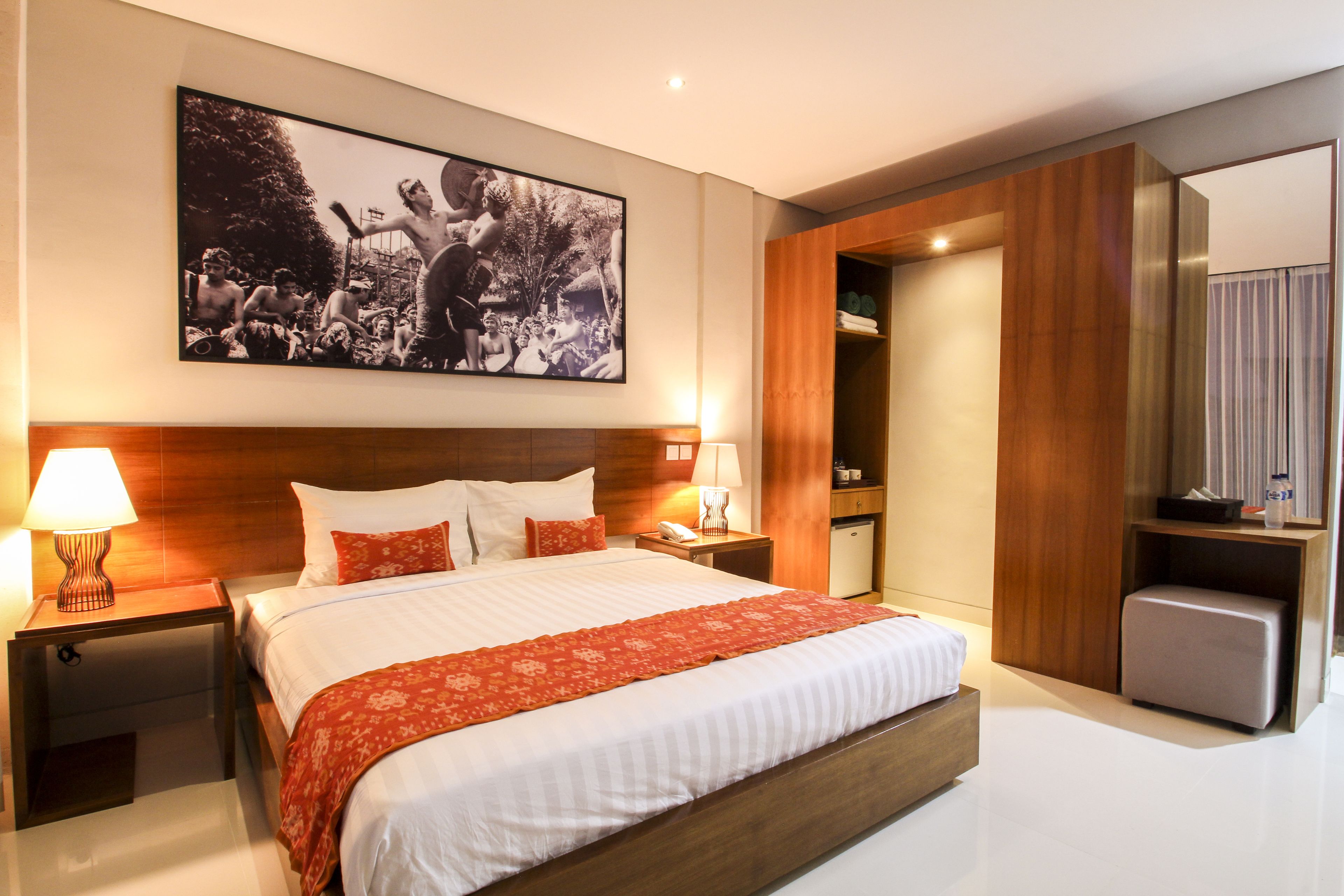 A modern hotel room with a large bed, wooden headboard, two bedside tables with lamps, a small seating area, and a large framed picture on the wall.