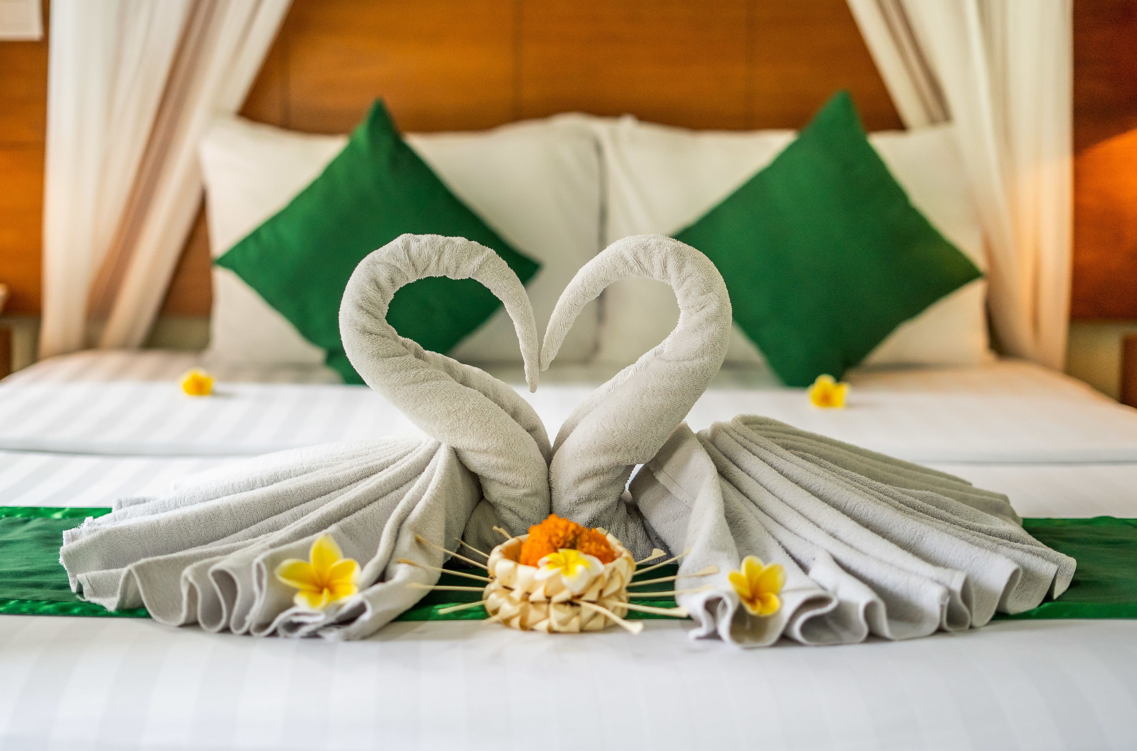 A neatly made bed with towels arranged in a heart shape, a small bowl with a yellow flower, and green pillows.