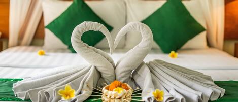 A neatly made bed with towels arranged in a heart shape, a small bowl with a yellow flower, and green pillows.