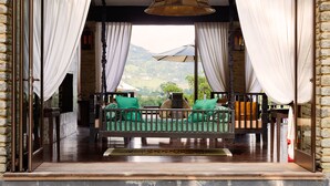 A wooden deck with a cushioned seating area, white curtains, and a view of a mountainous landscape. - The Pavilions Himalayas The Farm (Pokhara)