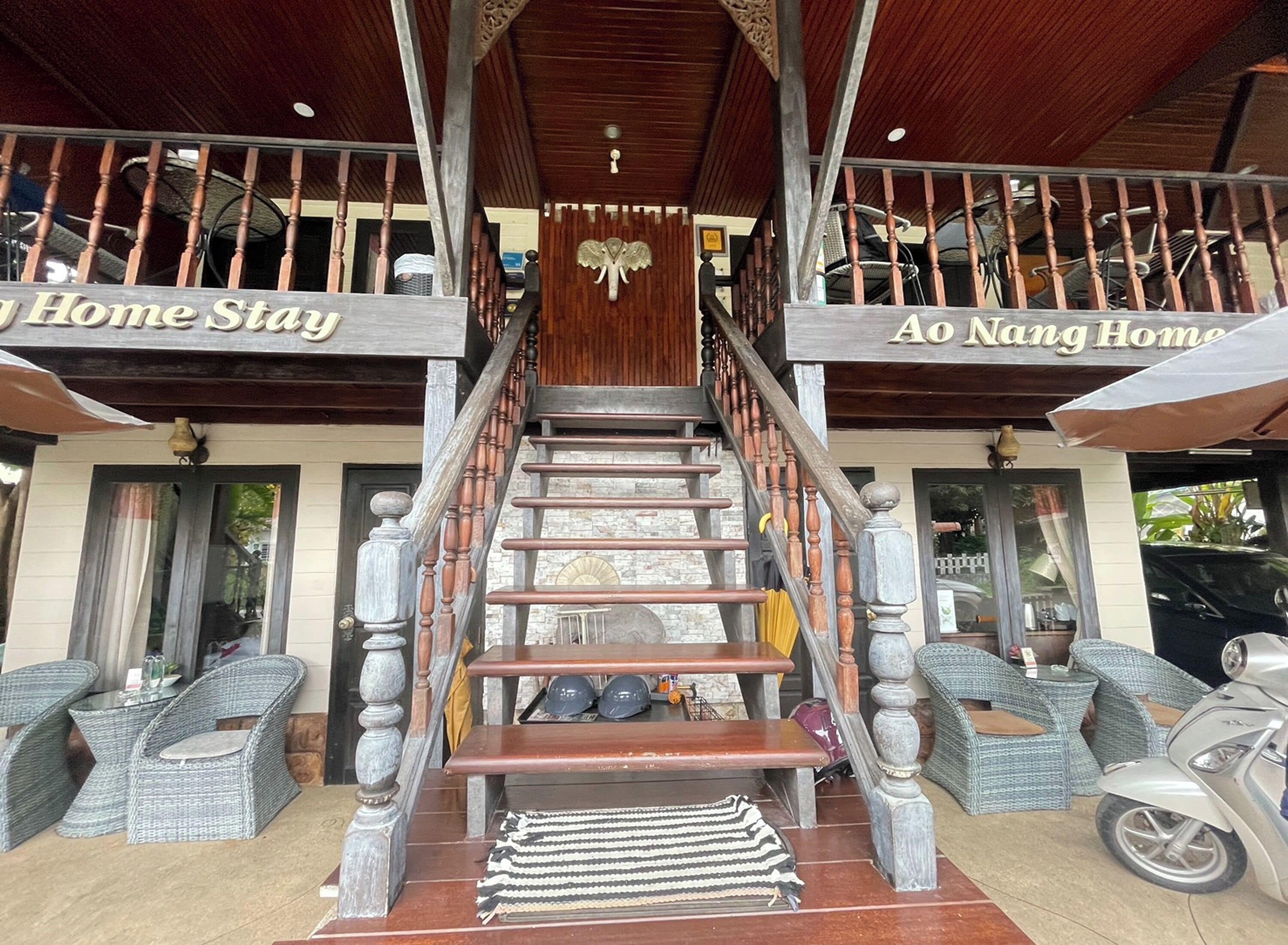 A staircase leading to a covered area with 'Home Stay' and 'Ao Nang Home' signs, wicker furniture, and a scooter.