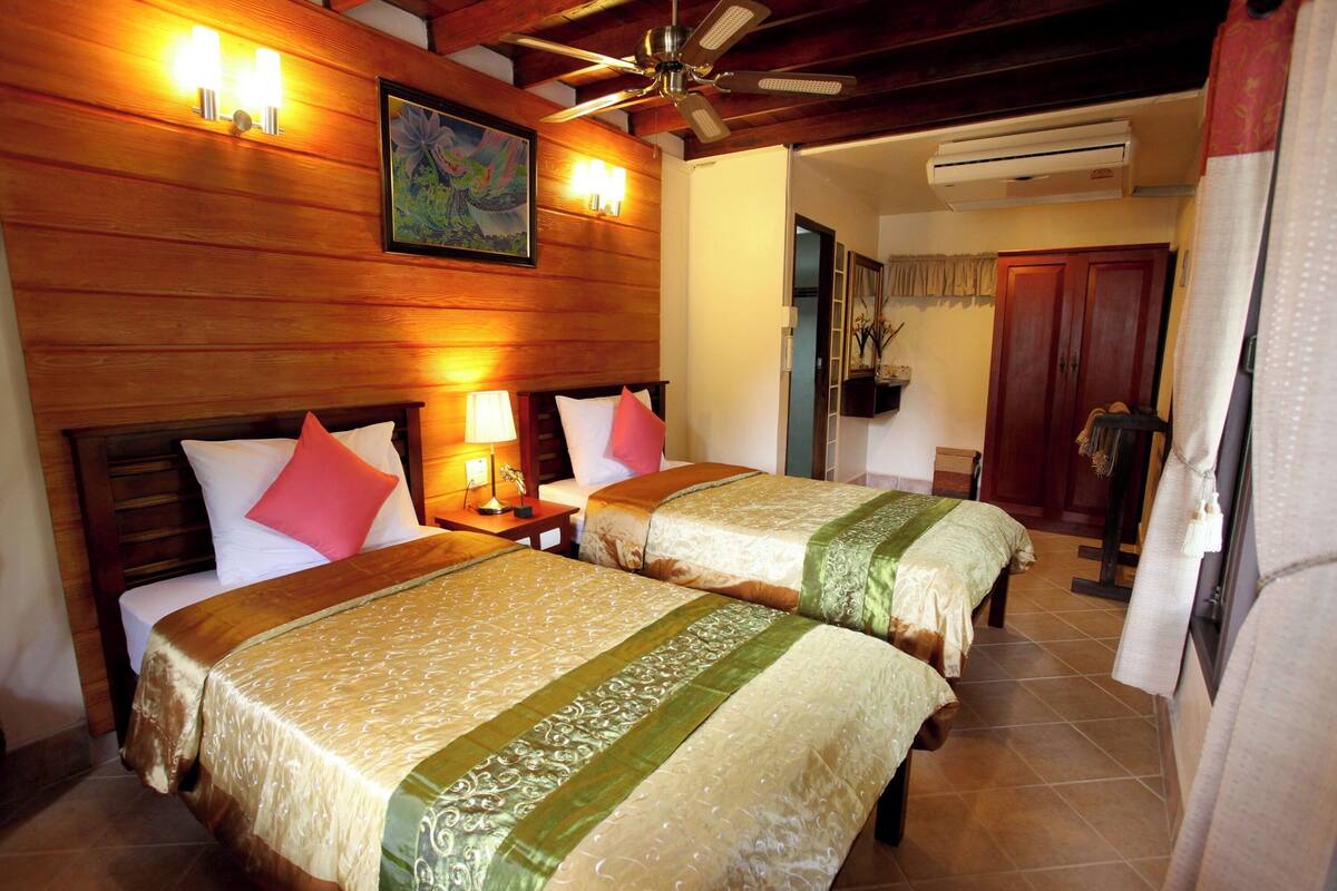 A hotel room with two beds, wooden headboards, a ceiling fan, and a painting on the wall.