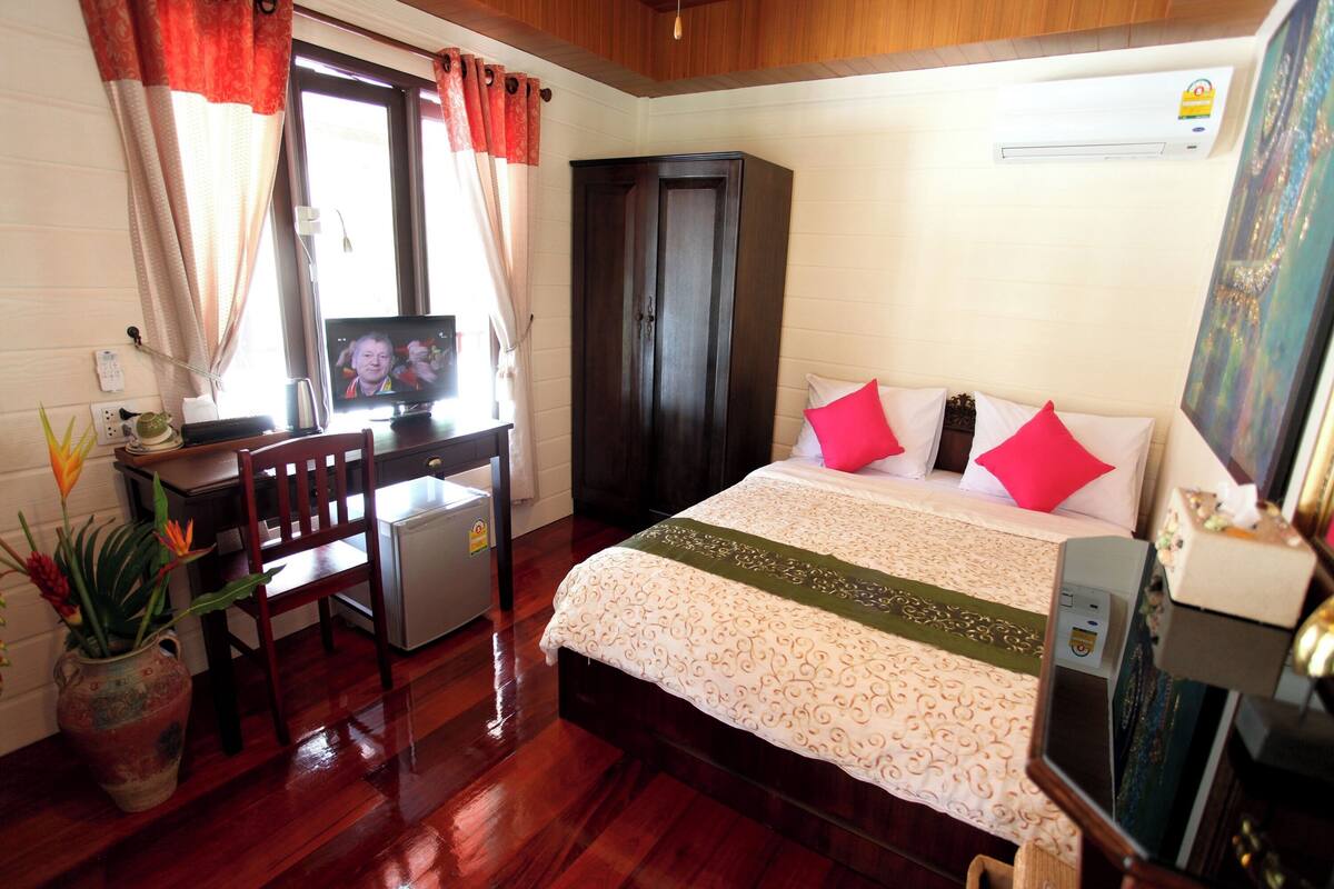 A hotel room with a bed, a desk with a television, a small refrigerator, a wardrobe, and a potted plant.