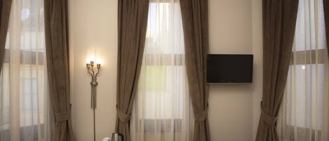 Una habitación de hotel con cama, escritorio con teléfono y cafetera, televisor montado en la pared y cortinas en las ventanas.