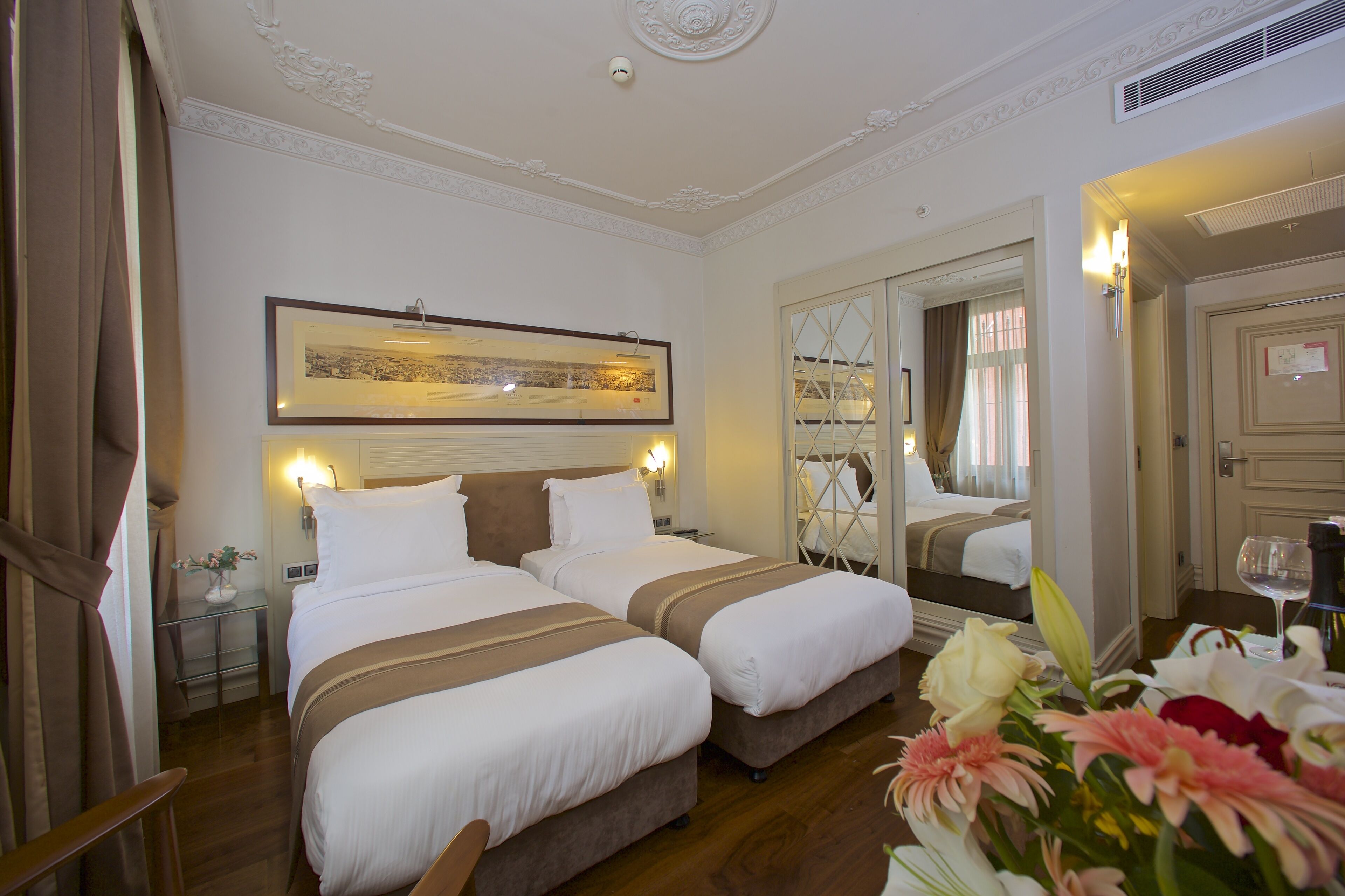 Une chambre d’hôtel avec deux lits, un grand miroir, un tableau encadré au mur et un vase de fleurs.