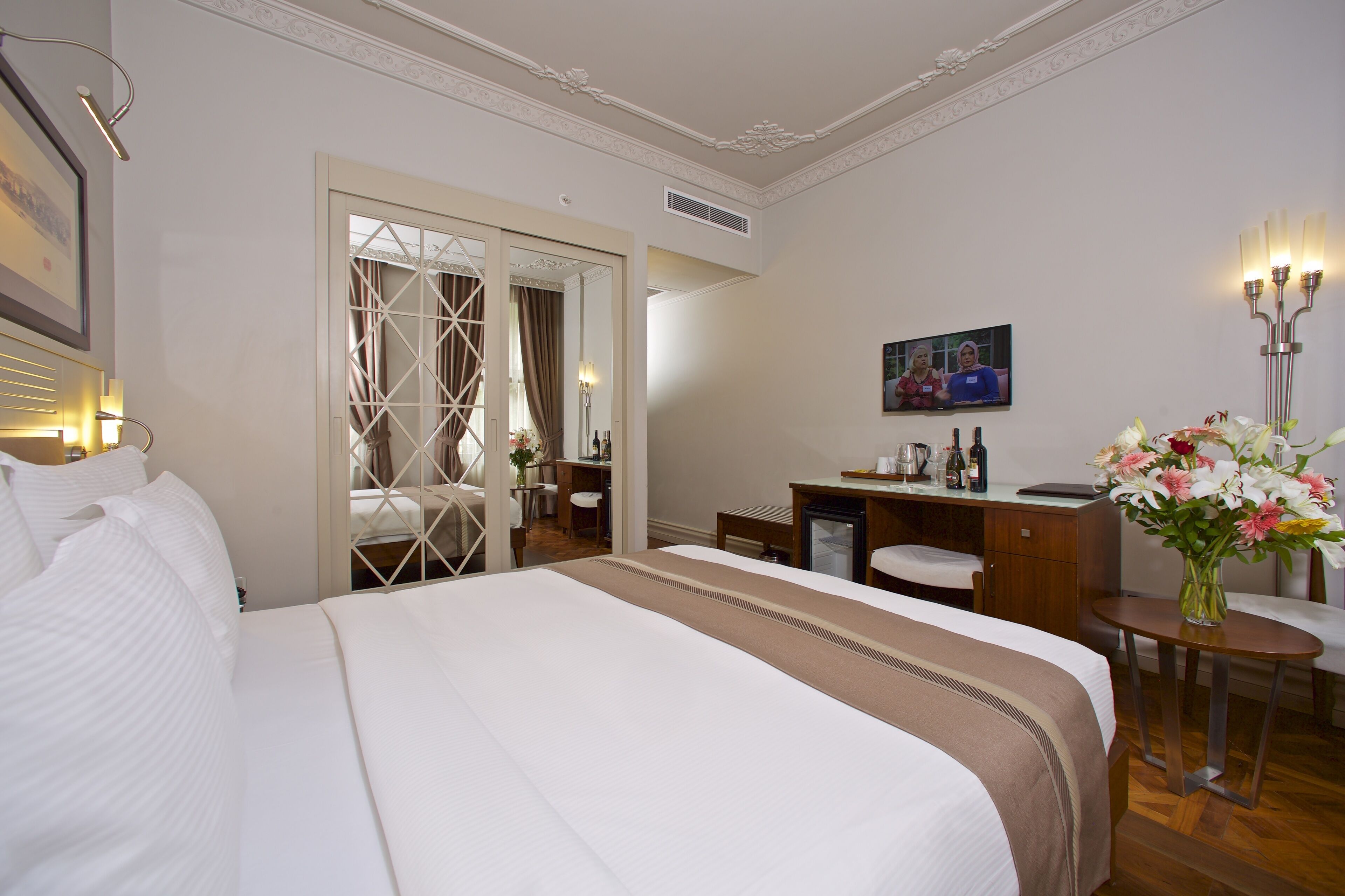 A hotel room with a large bed, a desk, a chair, a small table, a mirror, and a vase with flowers.