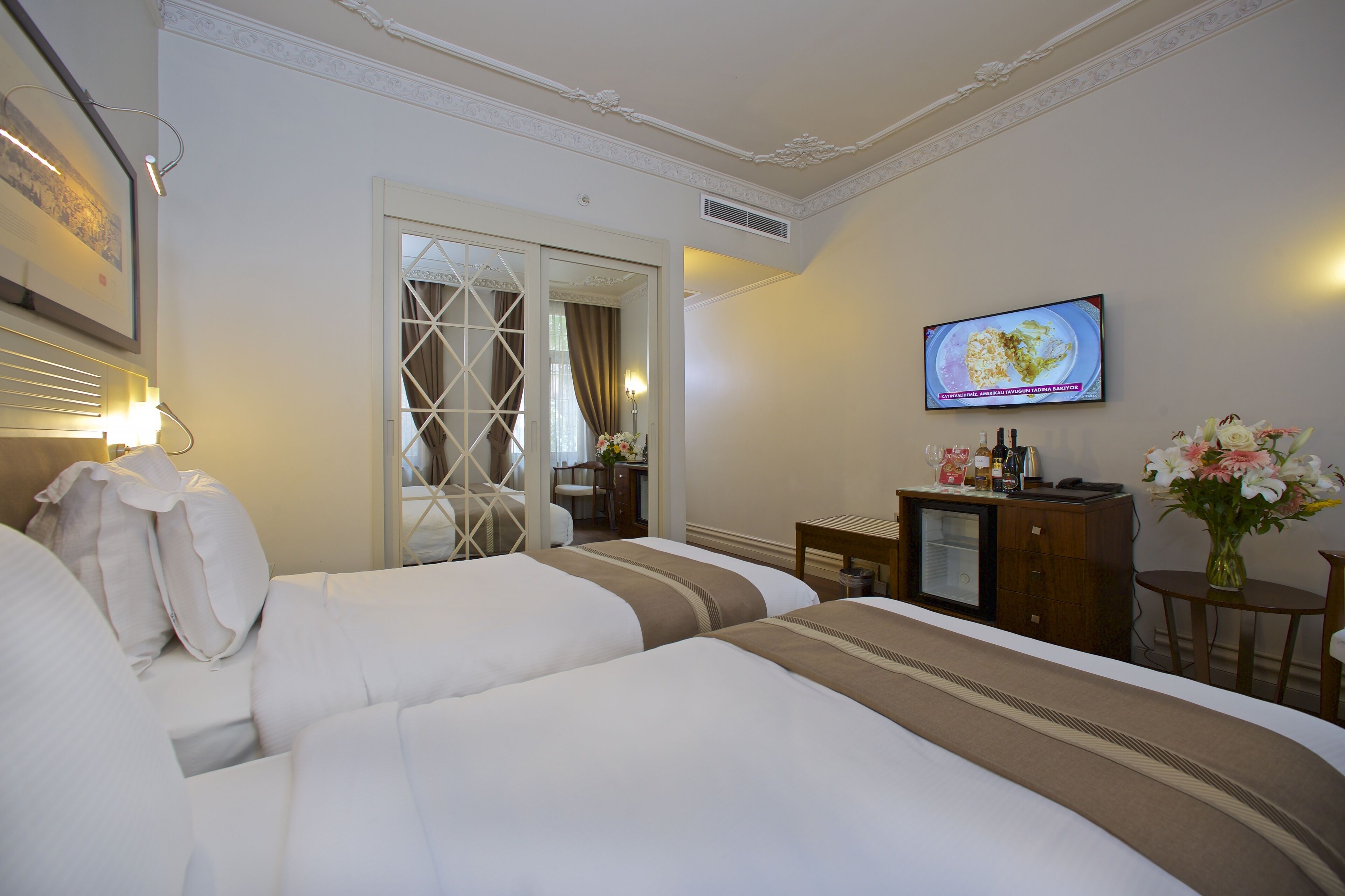 Une chambre d’hôtel avec deux lits, une télévision, un bureau avec un ordinateur, un petit réfrigérateur et un vase avec des fleurs.