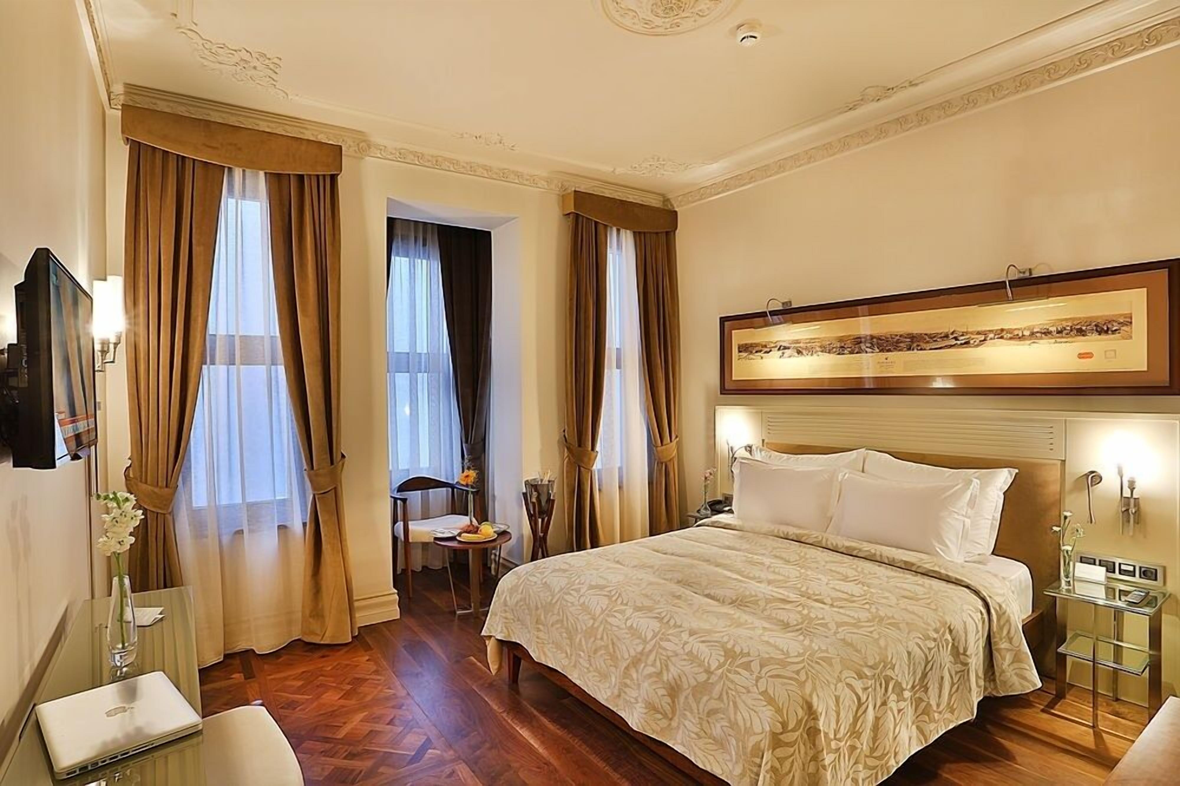 A hotel room with a large bed, a desk with a laptop, a chair, a small table with a vase, and a framed picture on the wall.